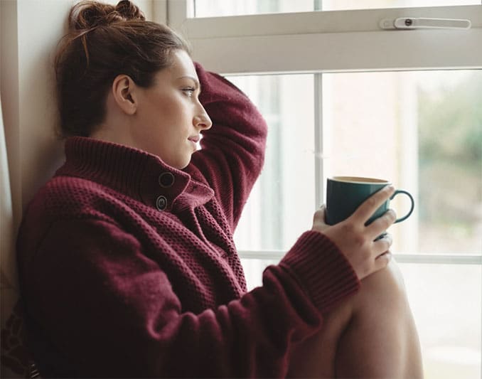 A mother in a quiet moment, sitting by a window holding a cup of tea, looking contemplative