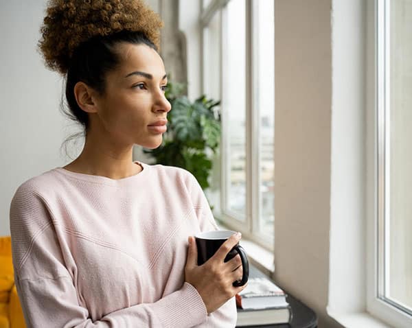 Pensive sad africanamerican woman standing at the window with a cup of coffee in her hands